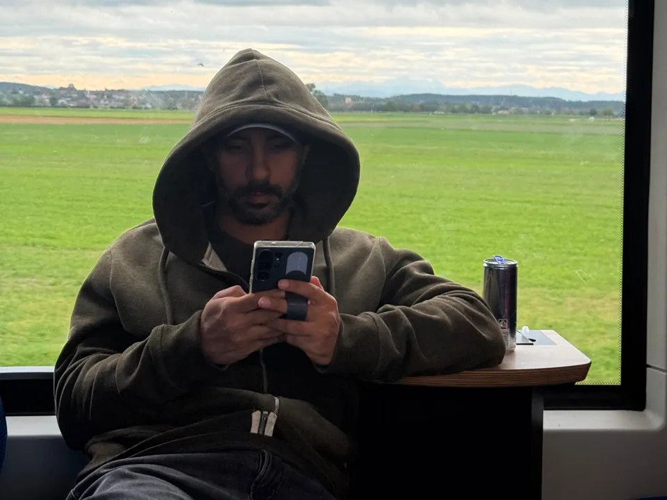 Sid in a hoodie on a train, looking at his phone, with green fields and hills outside the window